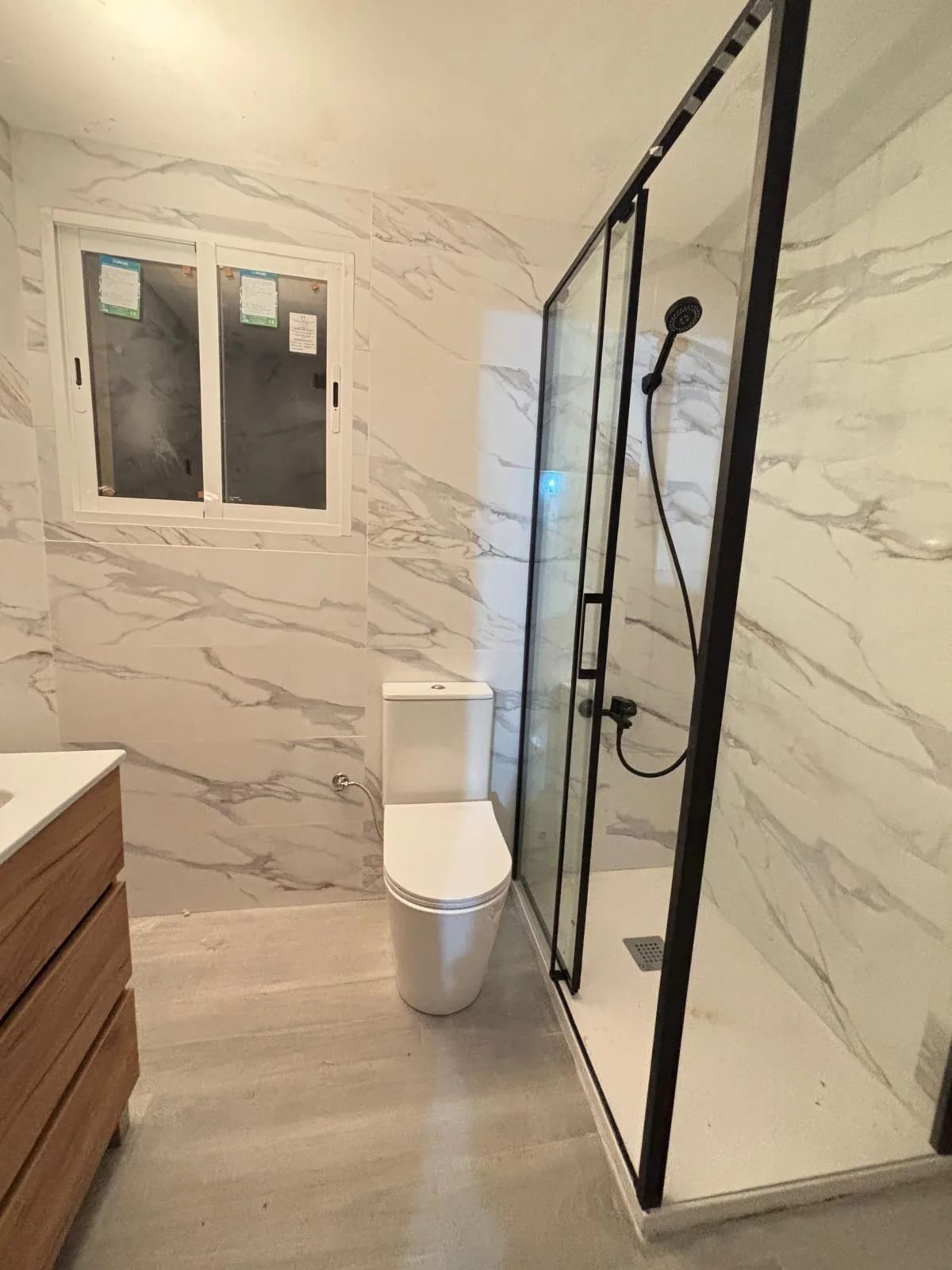 Baño reformado con ducha de mampara negra, inodoro blanco y paredes de mármol