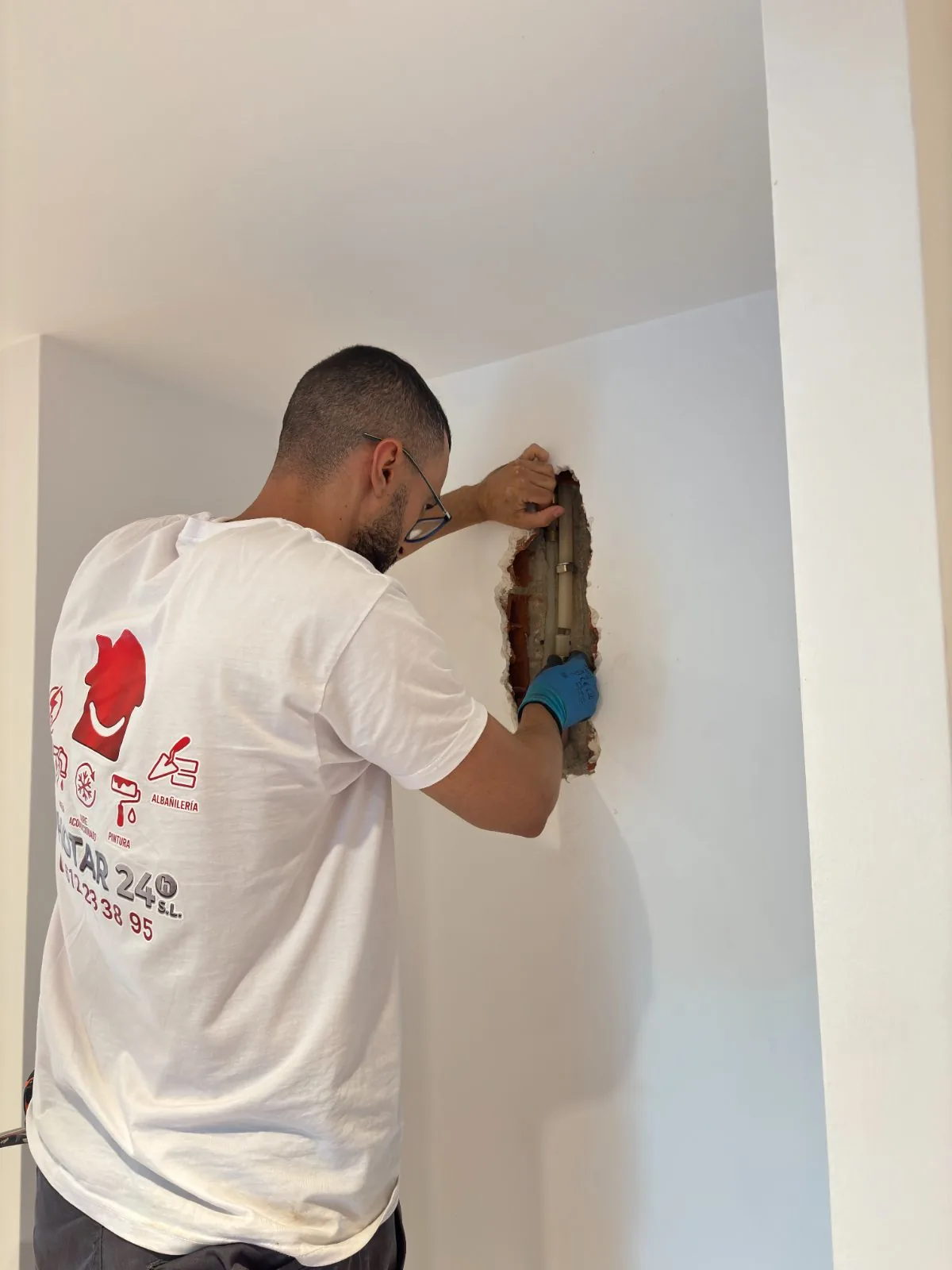 Fontanero con guantes azules trabajando en una tubería dentro de un hueco abierto en la pared